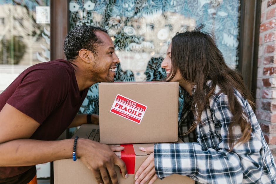 A man and woman outdoors during a home relocation process, smiling and facing each other while holding stacked cardboard boxes. The man, wearing a maroon t-shirt and a black wristband, is leaning forw