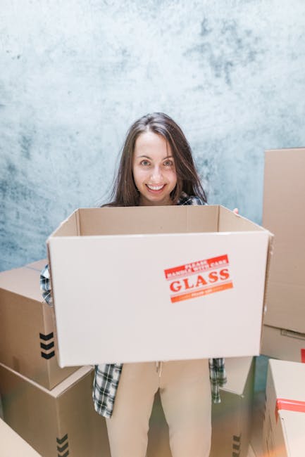 A man and woman outdoors during a home relocation process, smiling and facing each other while holding stacked cardboard boxes. The man, wearing a maroon t-shirt and a black wristband, is leaning forw