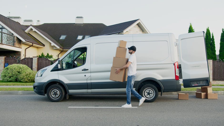 A man wearing a white t-shirt, blue jeans, and a black cap is loading three large cardboard boxes into the open rear cargo area of a white commercial van parked on a suburban street. The van is positioned parallel to the curb outside a residential property with a modern house, which features a dark roof, light-colored walls, and a wooden fence. Several smaller cardboard boxes are placed on the pavement near the van's rear, and the man is holding one of the boxes while walking towards the open van door, during daylight with clear weather. This scene depicts the process of house relocation, furniture transport, and packing and moving activities, with the focus on careful loading procedures aligned with professional removals services such as those provided by Man With a Van Eastcote.