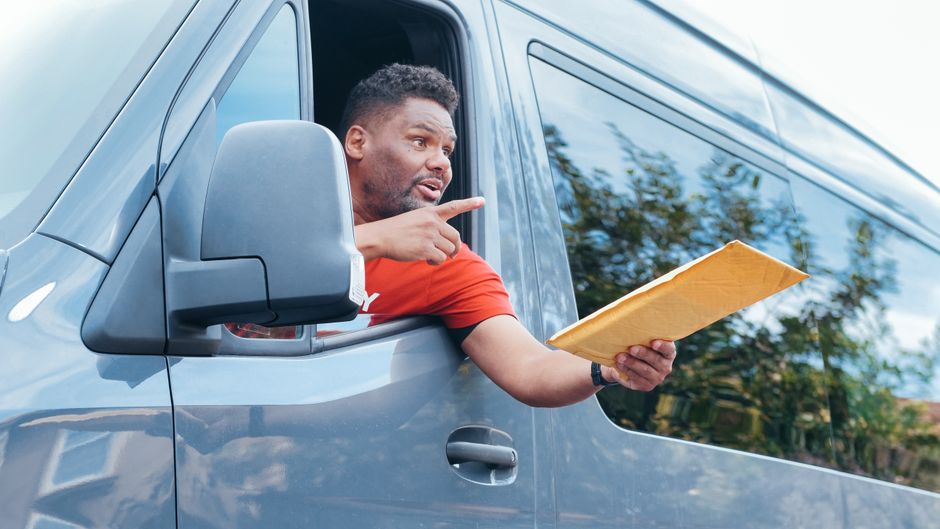 A man with a dark beard and curly hair, dressed in a red t-shirt, is leaning out of the open passenger window of a grey van, holding a large padded envelope or parcel wrapped in brown paper. He is pointing with his right hand and appears to be giving instructions or communicating during a home relocation or furniture transport process. The van is parked outdoors on a paved area, with a reflection of trees and blue sky visible on the side window. The background includes a blurred view of greenery and a residential property, indicating a suburban environment. This scene illustrates packing and loading activities associated with house removals, emphasizing the logistical aspect of moving services provided by companies such as Man With a Van Eastcoté.