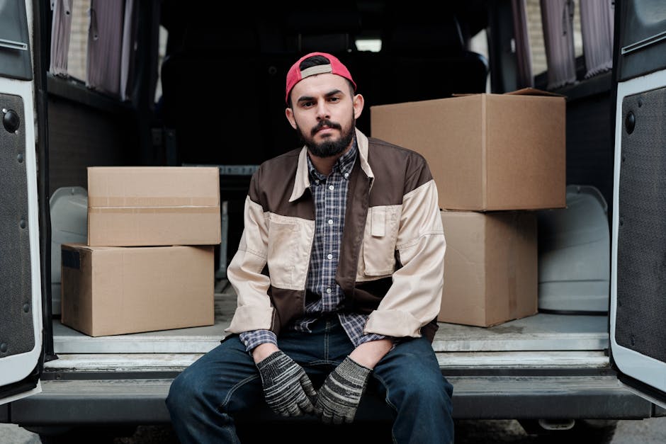 A man wearing a white t-shirt, blue jeans, and a black cap is loading three large cardboard boxes into the open rear cargo area of a white commercial van parked on a suburban street. The van is positioned parallel to the curb outside a residential property with a modern house, which features a dark roof, light-colored walls, and a wooden fence. Several smaller cardboard boxes are placed on the pavement near the van's rear, and the man is holding one of the boxes while walking towards the open van door, during daylight with clear weather. This scene depicts the process of house relocation, furniture transport, and packing and moving activities, with the focus on careful loading procedures aligned with professional removals services such as those provided by Man With a Van Eastcote.