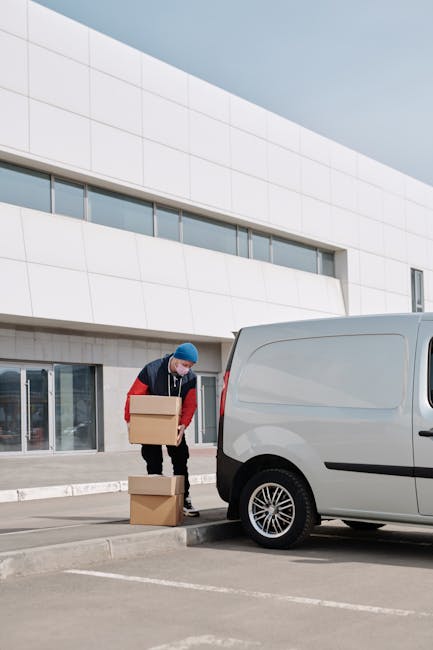 A man wearing a white polo shirt and a black cap is inside the rear of a large moving van, surrounded by neatly stacked cardboard boxes of various sizes, some open with flaps folded back. The boxes ar