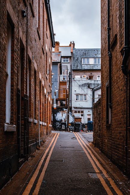 A narrow alleyway between two brick residential buildings during daytime, with double yellow lines along the asphalt pavement, leading to the back of a property. In the distance, there are several multi-storey buildings with grey facades, windows, and a wooden staircase descending from an upper level. Black trash bins and wheelie bins are positioned at the end of the alley towards a fenced yard area, indicating a typical urban environment. The scene appears to be part of a house relocation process on Eastcote High Road, with the setting suitable for furniture transport and packing and moving activities for local removals by Man With a Van Eastcote, as implied by the context of house removals through narrow streets and tight spaces.