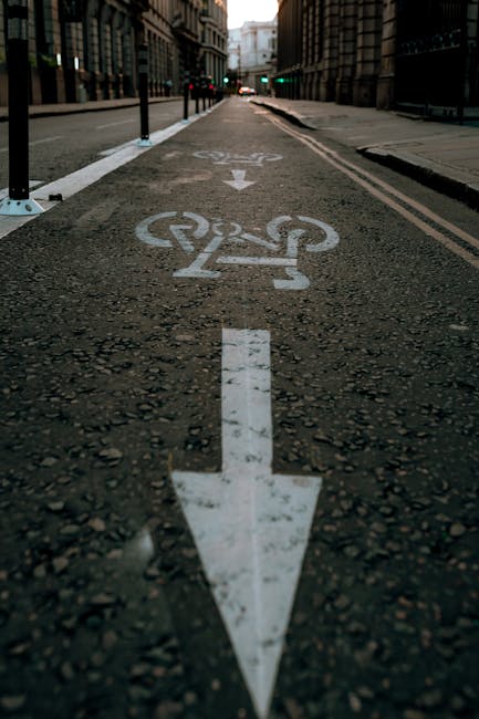 A paved city street with white bicycle lane markings, including a large downward arrow and a bicycle symbol, stretching into the distance beneath tall office buildings on both sides. The road is lined with black bollards and white lane dividers. In the background, traffic lights and distant buildings are visible under overcast sky lighting. This urban scene showcases the infrastructure for bicycle transport within a city environment, related to urban mobility and transportation planning, which may be relevant to house removals or moving services in an urban setting, as provided by Man With a Van Eastcote.