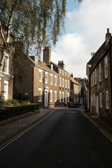 A narrow alleyway between two brick residential buildings during daytime, with double yellow lines along the asphalt pavement, leading to the back of a property. In the distance, there are several multi-storey buildings with grey facades, windows, and a wooden staircase descending from an upper level. Black trash bins and wheelie bins are positioned at the end of the alley towards a fenced yard area, indicating a typical urban environment. The scene appears to be part of a house relocation process on Eastcote High Road, with the setting suitable for furniture transport and packing and moving activities for local removals by Man With a Van Eastcote, as implied by the context of house removals through narrow streets and tight spaces.