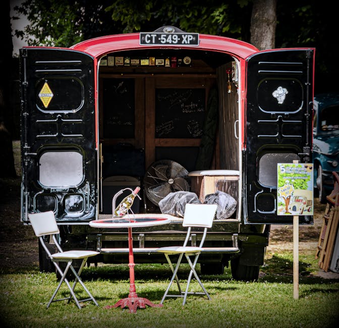The image shows a black and red vintage-style van with its rear doors open, revealing packaging materials, cardboard boxes, and furniture inside, positioned in an outdoor area surrounded by trees. In front of the van, there is a small round table with a white plastic bag and a plastic bottle, accompanied by two white folding chairs arranged on a grassy surface. A portable fan and items covered with plastic wrap are visible inside the van, indicating preparations for a home relocation or furniture transport. To the right of the van, a standing sign displays a vintage van illustration and the number '5', possibly indicating a promotional or informational display related to moving services. The setting appears to be part of a moving or clearance event, with natural lighting highlighting the scene, which illustrates the loading process as part of house removals by [COMPANY_NAME], supporting packing and transport logistics for local house moves.