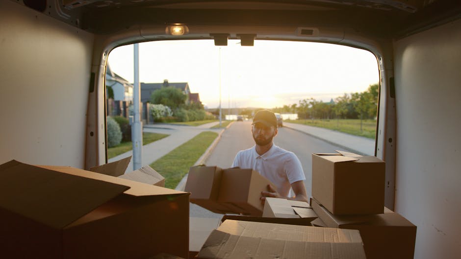 The image shows a black and red vintage-style van with its rear doors open, revealing packaging materials, cardboard boxes, and furniture inside, positioned in an outdoor area surrounded by trees. In front of the van, there is a small round table with a white plastic bag and a plastic bottle, accompanied by two white folding chairs arranged on a grassy surface. A portable fan and items covered with plastic wrap are visible inside the van, indicating preparations for a home relocation or furniture transport. To the right of the van, a standing sign displays a vintage van illustration and the number '5', possibly indicating a promotional or informational display related to moving services. The setting appears to be part of a moving or clearance event, with natural lighting highlighting the scene, which illustrates the loading process as part of house removals by [COMPANY_NAME], supporting packing and transport logistics for local house moves.