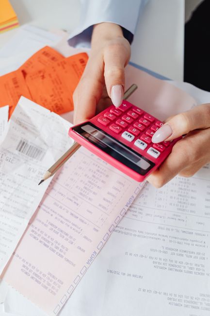 Close-up of a person's hand holding a pink calculator, with the hand resting on a collection of documents, receipts, and invoices spread out on a flat surface. The documents are partially visible, with some printed text and checkboxes, alongside a wooden pencil placed among the papers. In the background, there is an orange receipt or invoice and other paperwork, suggesting the process of cost calculation or financial planning related to house removals or relocation services. The scene is well-lit, emphasizing the detailed view of the paperwork and calculator, which are tools often used in managing moving logistics and home relocation costs. This image reflects the detailed planning phase involved in furniture transport and packing processes, as handled by professional removals services like Man With a Van Eastcote.