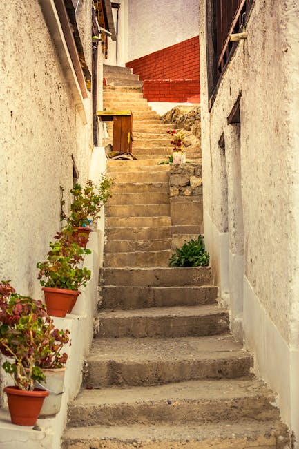 A narrow outdoor stone staircase with visible signs of age and wear, leading upwards between two white-painted walls of a residential property. On the left side of the stairs, there are several terracotta pots containing flowering plants and green foliage placed on small ledges attached to the wall. The stairs are uneven and slightly chipped, with some small plants growing in cracks. At the top of the staircase, wooden and brick structures are visible, possibly part of a garden or backyard area. The environment appears well-lit with natural daylight, highlighting the textures of the stone steps, potted plants, and surrounding walls. This setting illustrates typical vertical access in homes requiring careful furniture transport and home relocation planning, as managed by Man With a Van Eastcote.