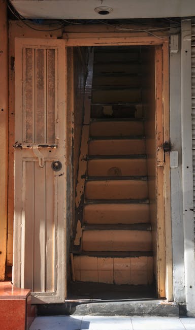 A narrow wooden staircase inside a property, with visible steps that are slightly worn and uneven, leading upward into the upper floors. The staircase is framed by unfinished wooden wall panels and a simple door on the left side, which appears to be part of a storage cupboard or small room. The surrounding wall structure shows exposed wood and insulation, indicating ongoing renovation or construction work. Natural light enters from the top, illuminating the steps and features, with shadows cast along the sides. The environment suggests a home undergoing a move or renovation, with potential challenges related to tight space access for furniture or packing materials, a situation often encountered during house relocations involving narrow staircases, as addressed by Man With a Van Eastcote's removals services.