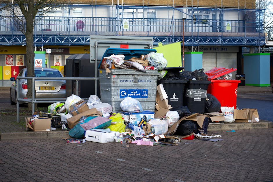 A collection of overflowing waste bins and scattered rubbish including cardboard boxes, plastic bags, paper and packaging materials situated beside a parking area on a paved surface. The bins include