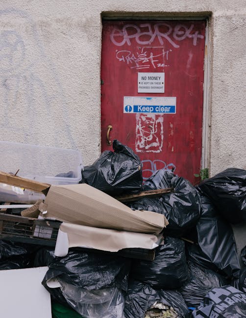 A collection of overflowing waste bins and scattered rubbish including cardboard boxes, plastic bags, paper and packaging materials situated beside a parking area on a paved surface. The bins include