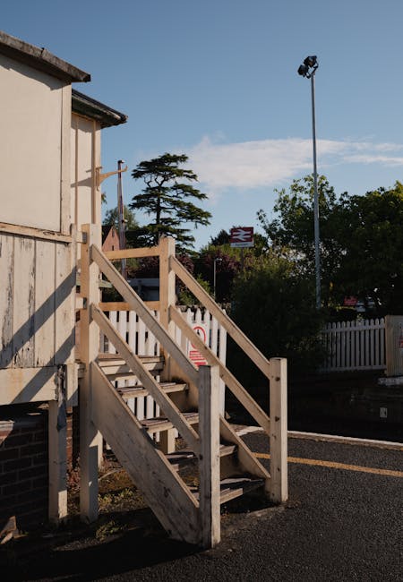 A wooden staircase with handrails and steps made of untreated timber leading up to a small access door attached to a building with exposed brickwork and weathered wooden cladding. The staircase is positioned outdoors on a paved surface, with a white picket fence, trees, and a lamppost visible in the background under a clear sky. The image captures the exterior environment during daytime, illustrating a typical example of tight access points that may require careful planning during home relocation or furniture transport, as handled by services like Man With a Van Eastcote.