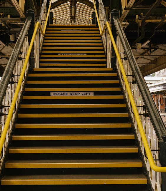 A set of industrial stairs inside a transportation hub or warehouse, with black and yellow non-slip treads and yellow safety railings on both sides. The stairs lead upwards toward an enclosed area with a curved wooden or metal structure overhead. Visible beneath the stairs are parts of metallic support beams and pipes, indicating a utilitarian environment. A white sticker on one of the middle steps displays the message 'PLEASE KEEP LEFT,' suggesting guidance for managing foot traffic during home relocation or furniture transport operations. The lighting appears to be natural or diffuse, creating minimal shadows and highlighting the functional nature of the staircase. The image captures a scene typical of loading or unloading zones involved in professional removals, as handled by companies like Man With a Van Eastcote.