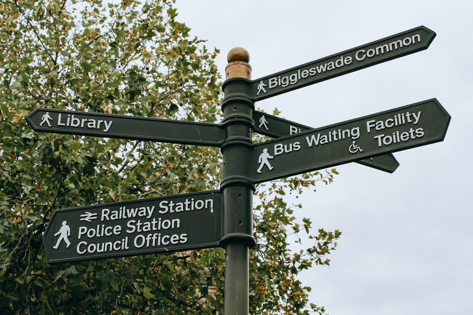A black public signpost with white lettering and icons, located outdoors amidst green leafy trees, providing directional guidance to various local amenities including Biggleswade Common, the library, railway station, police station, council offices, bus waiting facility, and toilets. The signpost features multiple directional arms, each pointing towards a different destination, with some arms displaying walking pedestrian icons for footpath routes. The surrounding environment has a cloudy sky, and the signpost appears to be positioned at a pavement or open public space, serving as a reference point for residents and visitors. This type of signage may be relevant in the context of home relocation or moving services, as it highlights key local points of interest and transit options near residential areas managed by companies like Man With a Van Eastcotе.