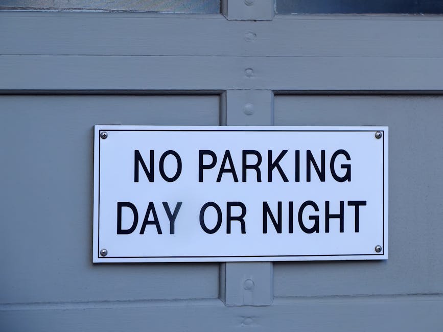 A rectangular white metal sign mounted on a light grey wooden panel indicates 'NO PARKING DAY OR NIGHT' in bold black uppercase letters. The sign is secured with four screws, one at each corner. The background includes visible horizontal and vertical wooden planks painted in grey, with a small metal bracket beneath the sign. The setting suggests outdoor environment near a property entrance where house removals or furniture transport may occur, potentially affecting parking regulations during home relocation or packing and moving activities, as managed by Man With a Van Eastcotе. The lighting is neutral, and the sign's design emphasizes clarity and visibility for drivers and pedestrians.