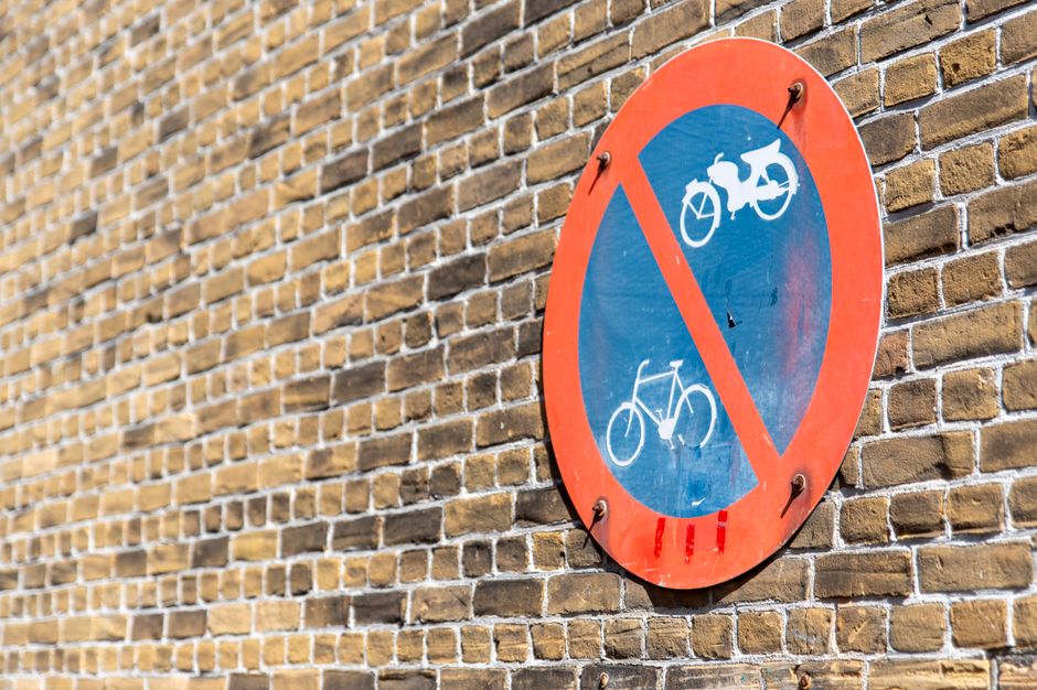 A round, red-bordered sign with a blue background is mounted on a brick wall, indicating that bicycle parking or cycling is not permitted in this area. The sign features two white bicycle icons with a diagonal red line crossing both of them, signaling a restriction on bikes. The brick wall behind the sign is composed of yellowish-brown bricks with visible mortar lines, creating a textured surface. The sign is secured with metal bolts at four points, and it appears to be outdoors, possibly on a street or in a residential area associated with house removals and moving services. The lighting suggests natural daylight, illuminating the wall and sign evenly. This signage is relevant to the logistics of house relocation, as it pertains to local traffic and parking restrictions that may affect the loading process for moving companies like Man With a Van Eastcote, especially when planning furniture transport or home relocation in Eastcote.