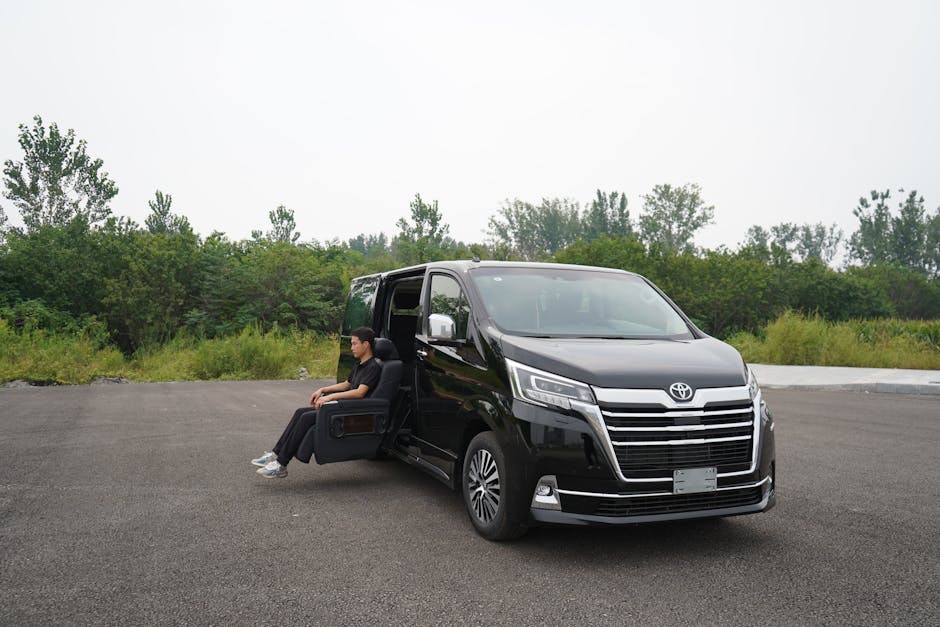 A man seated in a plush black armchair is positioned outside next to a black Toyota van, parked on an asphalt surface with a backdrop of green trees and shrubs under an overcast sky. The van's sliding