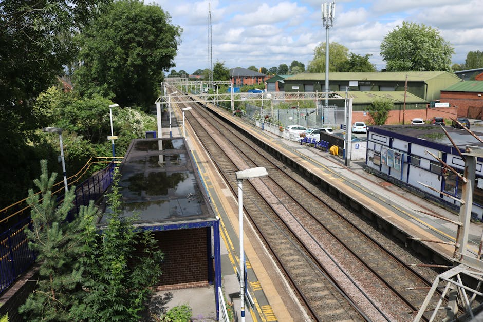 Image of Rayners Lane station platform in Eastcote during daytime, showing a wide view of the pedestrian walkway parallel to the railway tracks. The platform is paved with asphalt and has a yellow tac