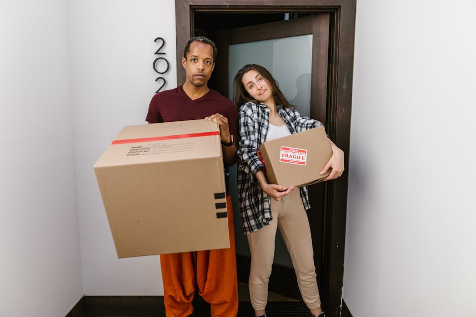 A man and a woman standing inside a doorway, each holding cardboard boxes during a home relocation or packing process. The man, on the left, is wearing a maroon T-shirt and loose orange trousers, with a serious expression, and is holding a large box with red and black markings indicating fragile contents. The woman, on the right, is dressed in a checked shirt over a white T-shirt and beige trousers, smiling slightly, and holding a smaller box with a 'Fragile' label. The doorway behind them leads outside or to another room, with part of a dark wooden frame visible. The scene takes place indoors under natural or ambient lighting, with packing materials and boxes suggesting an ongoing moving or furniture transport operation managed by [COMPANY_NAME], a professional removals service specializing in house moves and furniture transport.