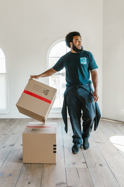 A man with an Afro hairstyle and a beard, wearing a blue t-shirt with a logo on the chest and dark trousers, is inside a bright room with white walls and large arched windows. He is holding a cardboard box with red tape, preparing to place it on top of another box on the wooden floor. The room appears to be part of a house or apartment, with natural light coming through the window, and the scene depicts packing or furniture transport as part of a home relocation process. This image is associated with professional removals services by Man With a Van Eastcote, highlighting packing and loading activities during house moves or furniture transport.