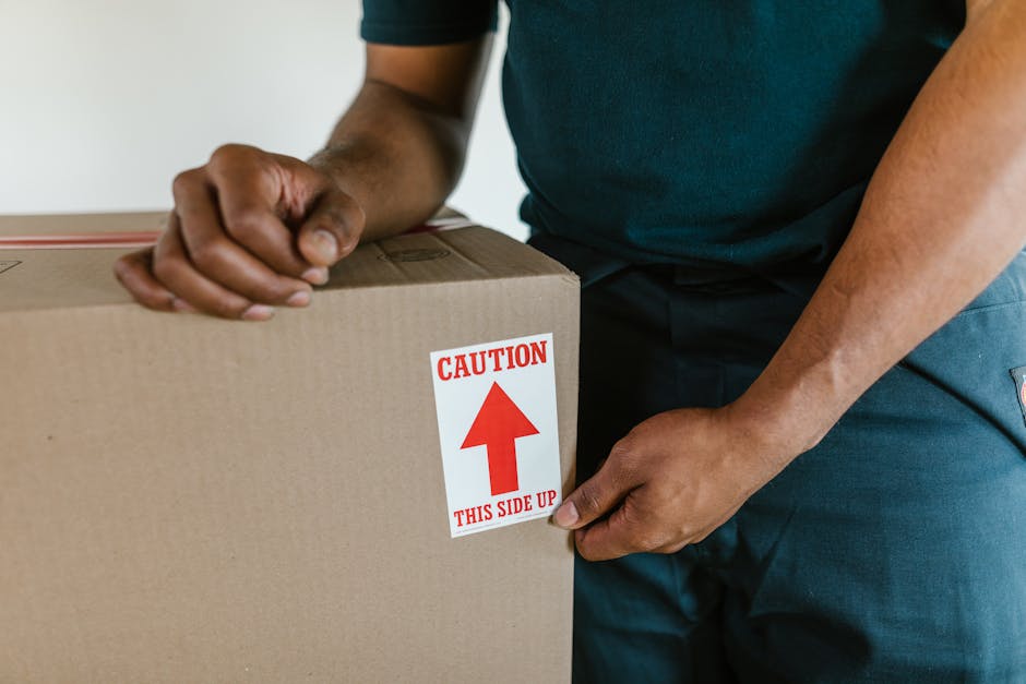 A man and a woman standing inside a doorway, each holding cardboard boxes during a home relocation or packing process. The man, on the left, is wearing a maroon T-shirt and loose orange trousers, with a serious expression, and is holding a large box with red and black markings indicating fragile contents. The woman, on the right, is dressed in a checked shirt over a white T-shirt and beige trousers, smiling slightly, and holding a smaller box with a 'Fragile' label. The doorway behind them leads outside or to another room, with part of a dark wooden frame visible. The scene takes place indoors under natural or ambient lighting, with packing materials and boxes suggesting an ongoing moving or furniture transport operation managed by [COMPANY_NAME], a professional removals service specializing in house moves and furniture transport.